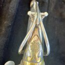 Detailní pohled na horní část saxofonu v stříbrném povrchu s mosaznými prvky. Na těle nástroje je vidět kulaté logo s nápisem H. SELMER PARIS. Povrch…