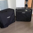 Černý kytarový kombozesilovač s nápisem MESA ENGINEERING na přední mřížce a EXPRESS na ovládacím panelu. Vedle zesilovače je černý ochranný kryt s bí…
