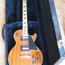 Elektrická kytara s trupem ve tvaru Les Paul v žlutohnatém plamenovém provedení, umístěná v černém tvrdém kufru. Na hlavě hmatníku je nápis Ovation a…