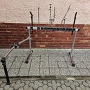 Kovový stojan pro perkuse s chromovaným povrchem a černými plastovými prvky. V horní části se nachází vodorovná lišta s řadou černých držáků a několi…