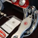 Detailní pohled na stříbrný kovový pedál s červenými gumovými hroty. Na těle pedálu i na hrotech je viditelné červené logo s písmenem G. Vedle pedálu…