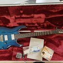 Modrá elektrická kytara s prožilkovaným povrchem v otevřeném kufru s červenou výstelkou. Na hlavě hmatníku je logo Ibanez. Kytara má dva humbuckery,…