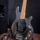 Elektrická basová kytara s tmavým třpytivým povrchem těla a světle hnědým hnedem hmatníkem. Nástroj má černý pickguard a chromované kovové části. Na…