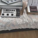 Na fotografii je bílý basový procesor s nápisem BOSS GT-1B BASS MULTI-FX PROCESSOR a logem COSM. Vedle přístroje je umístěna jeho originální krabice…