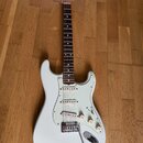 Bílá elektrická kytara typu Stratocaster s bílým pickguardem a hnedým hmatníkem. Na hlavě je vidět nápis Fender. Nástroj má tři snímače, tři ovládací…