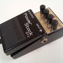 Černý efekt pedál s kovovým ovládacím panelem. Na horní části je bílý nápis BOSS a označení Power Stack ST-2 s šipkami OUTPUT a INPUT. Na panelu jsou…