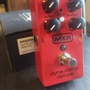 Červený efekt pedál s nápisem MXR a dyna comp deluxe. Na horní straně jsou čtyři černé ovládací knoflíky s popisky CLEAN, TONE, OUTPUT a SENSITIVITY,…