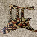 Kytarový pickguard s barevným geometrickým vzorem v hnědých, žlutých a červených odstínech. Má výřezy pro dva snímače a otvory pro šrouby a ovládací…