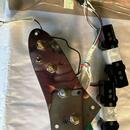 Chromovaný kovový pickguard s instalovanými potenciometry a přepínačem. Vedle něj jsou dva černé humbuckery s viditelným nápisem Maintenance. Kabely…