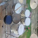 Celá bicí souprava v krémové barvě umístěná na dřevěné terase. Sestává z několika bubnů, dvou činelů značky MEINL LIVE SOUND a černého sedáku. Na jed…
