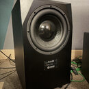 Černý studiový subwoofer s matným povrchem a hranatým tvarem. Na přední straně je umístěn velký kruhový reproduktor a pod ním štítek s nápisy „On Sub…