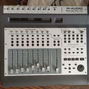 Šedý audio interface a kontrolní povrch s nápisem M-AUDIO PROJECTMIX I/O firewire audio interface | control surface. Horní panel obsahuje osm vstupní…