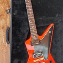 Oranžová elektrická kytara s hranatým tělem v podobě šipky, položená v černém kufru. Na hlavě hředu je zlaté logo Gibson. Kytara má dva humbuckery, z…