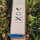 Stříbrný efekt pedál s rýhovaným povrchem a chromovanými hranami. Uprostřed je nápis VOX zopakovaný dvakrát, vlevo je svisle napsáno AMPLIFIER a vpra…
