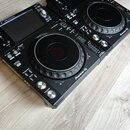 Dvě černé DJdecksy značky Pioneer DJ umístěné vedle sebe na světlé dřevěné podlaze. Každý přístroj má velký ovládací jog-wheel, displej a řadu tlačít…