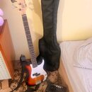 Elektrická basová kytara s dřevem v přirozené barvě, hnabodovaným korpusem v sunburst provedení a bílým pickguardem. Na hlavě je nápis "PHASE SERIES"…