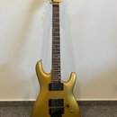 Zlatá elektrická kytara s tmavým hmatníkem a dvěma humbuckery. Na hlavě je viditelné logo s nápisem "Cort". Povrch nástroje je v dobrém stavu s metal…