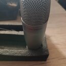 Stříbrný kondenzátorový mikrofon s kovovou síťkou v horní části. Na těle je nápis USB STUDIO CONDENSER. Mikrofon stojí v tmavě šedé pěnové vložce na…