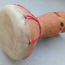 Djembe s tělem z hnědého dřeva a napnutou kůží v přirozené bílošedé barvě. Membrána je upevněna červeným provázkem. Tělo nástroje je v horní části zd…