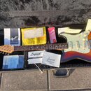 Elektrická kytara s tmeňem v sunburst provedení, bílým pickguardem a javorovým hmatníkem leží v černém plišovém kufru. Na hlavě kytary je logo Fender…