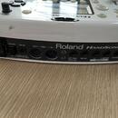 Bílé elektronické bicí zařízení s nápisem Roland HANDSONIC 10. Zadní panel obsahuje konektory POWER, DC IN, OUT — MIDI — IN, EXT TRIG FOOT SW, MIX IN…
