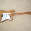 Elektrická kytara s přírodním dřevěným povrchem těla a hmatníkem. Má bílý pickguard, tři snímače a nápis "Cort" na hlavě hmatníku. Nástroj vykazuje z…