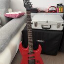 Červená elektrická kytara s černým hmatníkem a černou hlavou. Na hlavě je bílý nápis "Jet Rock". Kytara má dva snímače, ovládací knoflíky a je umístě…
