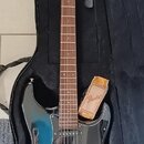 Modře přechodová elektrická kytara v černém obalu. Na hlavě hmatníku je viditelné logo Fender. Kytara má zlatý hardware, černý pickguard a v obalu ve…