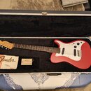 Červená elektrická kytara s bílým pickguardem v černém polstrovaném kufru. Na hlavě hmatníku je nápis Fender Stratocaster. V kufru vedle nástroje lež…