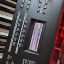 Detailní pohled na ovládací panel černé klávesové syntezátoru s displejem a řadou šedých tlačítek. Na displeji je vidět text „MOTIF“, „USR:001(001)“…