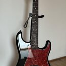 Černá elektrická kytara s červeným vzorovaným pickguardem a hnědým hmatníkem. Na hlavě nástroje je viditelné logo Squier. Kytara má jeden humbucker v…