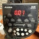 Černý elektronický modul Alesis DM6 DRUM MODULE s červeným digitálním displejem. Na panelu jsou různé ovládací tlačítka a nápisy jako TEMPO, KIT, VOI…