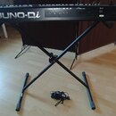 Černý digitální klavír s nápisem Roland JUNO-Di umístěný na černém X-stojanu. Vedle nástroje leží černý napájecí adaptér s kabelem. Povrch klavíru a…