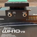 Fotografie zachycuje detail zadní strany černého wah pedálu a přiložený manuál. Na přístroji jsou vidět konektory s nápisy INPUT, DC9V a OUTPUT. Pod…