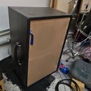 Černý hranatý reproduktorový box s rukojetí na boku. Zadní strana je z nebarveného dřeva nebo překližky, kde je připevněna modrá textilní páska s bíl…