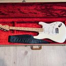 Bílá elektrická kytara typu Stratocaster s javorovým hmatníkem v otevřeném kufru s červeným vnitřním polstrováním. Na hlavě nástroje je viditelné log…