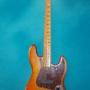 Elektrická basová kytara s přírodním dřevěným povrchem těla a hmatníkem. Na hlavě je viditelné logo Fender. Kytara má tmavě hnědý pickguard s mramoro…