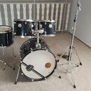 Černá bicí souprava s chromovaným kováním, která obsahuje basový buben, dva tom-tomy a jeden snare drum. Na přední straně basového bubnu je bílý kože…