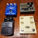 Čtyři efektové pedály umístěné na dřevěném povrchu. Modrý pedal s nápisy DIGITAL DELAY, Harley Benton a True Bypass; zelený pedal s nápisy Easy Drive…