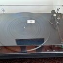Gramofon v průhledném plastovém krytu s černým podkladem. Na talíři je umístěna černá deska s bílým štítkem uprostřed. V pravém dolním rohu je nápis…