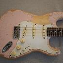 Elektrická kytara s růžovým povrchem, který vykazuje výrazné opotřebení a rozestřený lak v okrajích těla. Nástroj má bílý pickguard, tři snímače a tř…