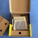 V otevřené kartonové krabici se nachází žlutý efekt pedál zabalený v průhledném plastovém obalu. Na horní části pedálu jsou vidět tři ovládací knoflí…