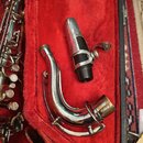 Na červeném povrchu pouzdra leží stříbrný kovový krk saxofonu a černý s kovovým povrchem mouthpiece. Na obou předmětech nejsou vidět žádné značky ani…