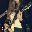 Žena s dlouhými hnědými vlasy drží v rukou elektrickou kytaru. Nástroj má krémové tělo s černým lemováním a hnědý hmatník. Na hlavě kytary jsou vidět…