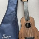 Hnědá ukulele s tmavým hmatníkem a modrý textilní obal. Na hlavě nástroje i na obalu je bílým písmem nápis Flight. Kolem ozvučovací otvory je vyřezáv…