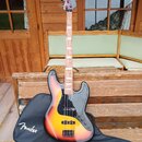 Elektrická basová kytara s trupem v sunburst provedení a černým pickguardem. Na hlavě hmatníku není žádný viditelný nápis, na černém obalu v pozadí j…