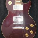 Tmavě červená elektrická kytara s tvarem Les Paul a krémovým lemováním. Na těle jsou dva humbuckery, čtyři zlaté ovládací knofíky a stoper s kovovým…