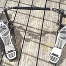 Dvojpedál na bicí buben v černo-stříbrném provedení s kovovými komponenty. Na obou šlapkách je viditelné logo MAPEX v oválném rámu. Povrch pedálů vyk…