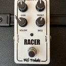 Bílý efekt pedál s nápisy RACER a MH pedals. Obsahuje čtyři černé ovládací knoflíky s popisky GAIN, TREBLE, VOLUME a BASS, vstup INPUT, výstup OUTPUT…