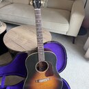 Akustická kytara s tmavě hnědým sunburst povrchem korpusu a tmavým hnedým hmatníkem. Na hlavě nástroje je bílý nápis Gibson. Kytara je umístěna v ote…